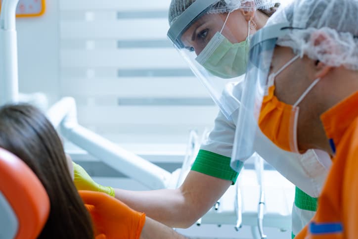 Dentist and hygienist treating patient in office, wearing PPE to protect against COVID-19