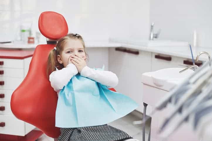 Frightened little girl experiences dental anxiety at dentist office, covers mouth with hands