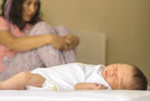 Newborn infant sleeping on bed while anxious, depressed mother sits nearby hugging her knees to her chest
