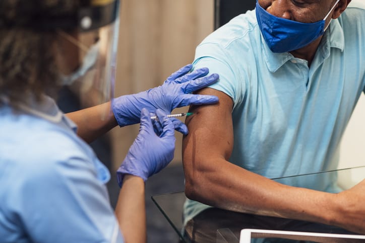 A mixed race female nurse wearing a protective face shield, surgical mask and protective gloves administering the COVID-19 vaccine to a senior black man.