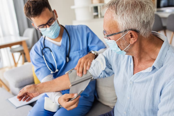 Male ambulatory care nurse measures blood pressure to senior man with mask during home visit