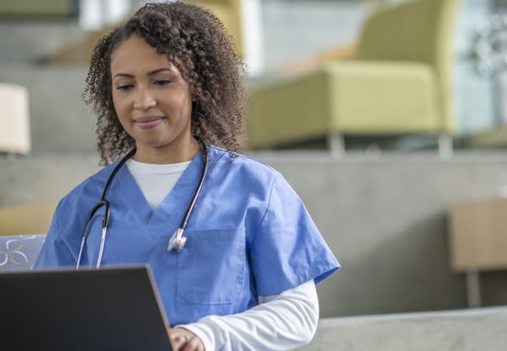 Nursing professional in scrubs happily attends online webinar on laptop