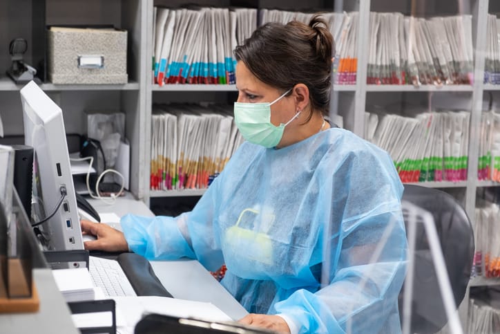 Receptionist at dental office wearing mask and gown for protection during the Covid-19 pandemic. She is working at the computer with patient files in the background.