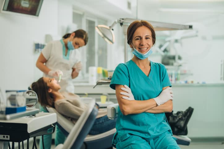 Smiling dentist sitting at dentist's office with her arms crossed and looking at camera, with a staff member treating a patient in the background.