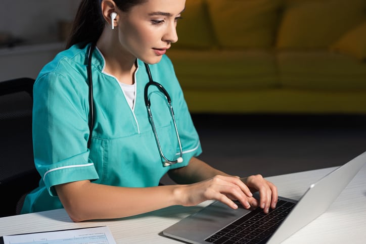 Nurse in uniform sitting at table and using laptop to watch webinar
