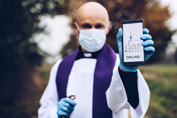 Priest in a medical mask and protective gloves holds a mobile phone with information about online meetings during COVID-19 pandemic.