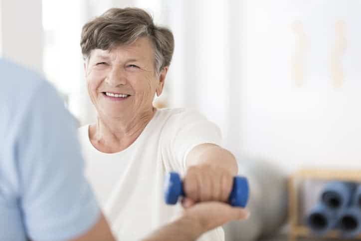 Happy senior woman exercising arms with small weights with help from a physical therapist. Stroke hemiplegia concept.