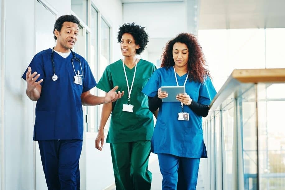 Male and female nurses in scrubs walk through hospital, talking together, one is using digital tablet