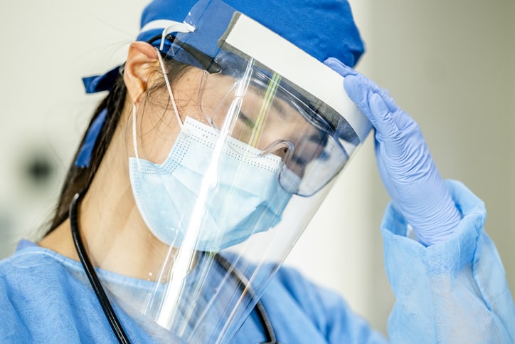 Asian female doctor appears to be stressed. She stands with hand on forehead, wearing safety goggles, face mask, surgical cap and face shield. Nursing burnout concept.
