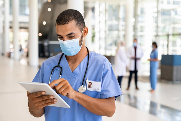 Young male nurse using digital tablet in corridor while wearing surgical mask.