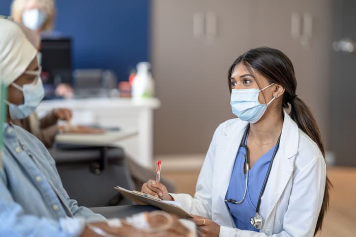 A female doctor talks with a cancer patient who is receiving treatment, and both are wearing medical masks to protect them from COVID-19.