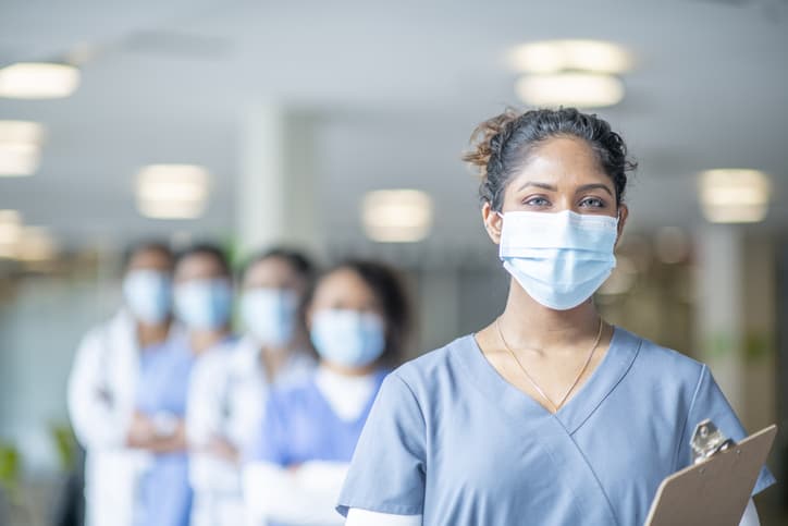 Female doctor or nurse leader wearing scrubs and a face mask stands in front of her colleagues at the hospital. Healthcare leadership concept.