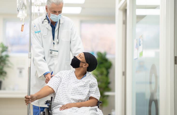 A male doctor is helping push his cancer patient on a wheelchair back to her hospital room, both wearing face masks during COVID-19 pandemic, preventing neutropenia concept.
