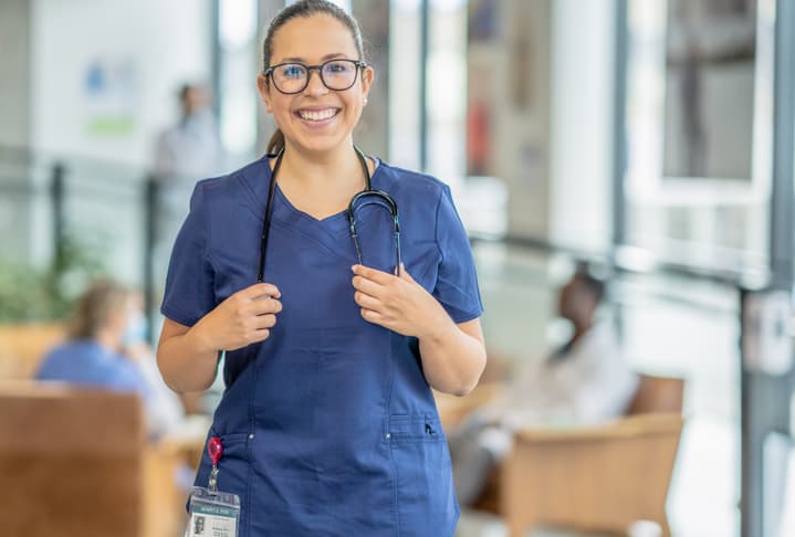 A portrait of a friendly and smiling nurse practitioner of Latin and Hispanic descent wearing blue medical scrubs.