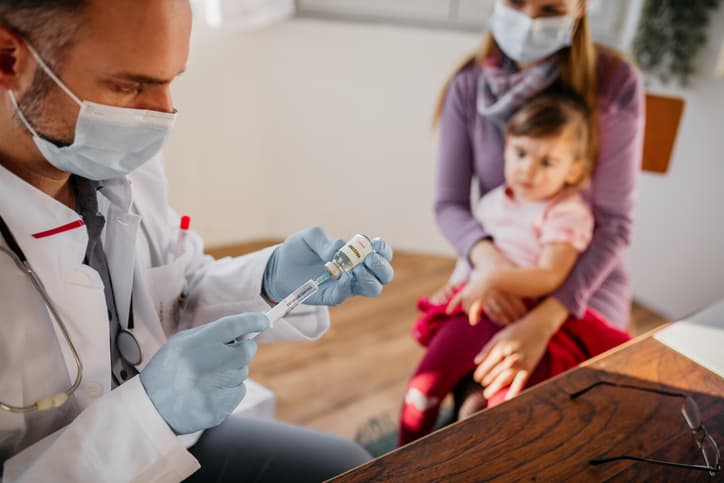 Doctor wearing mask and gloves prepares COVID-19 vaccine for toddler girl in mother's lap